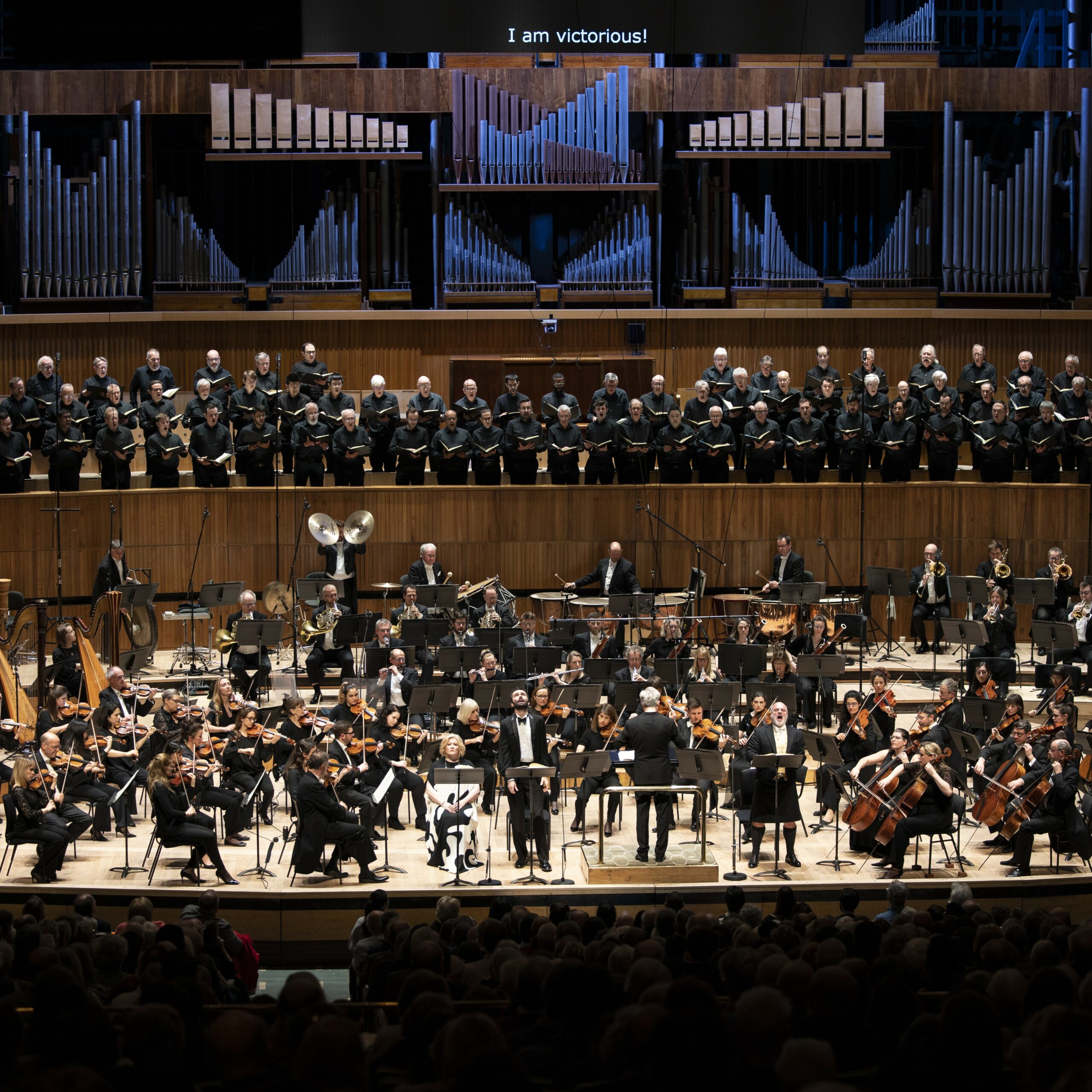 A choir, orchestra, soloists and conductor on stage at the Royal Festival Hall, wearing black.