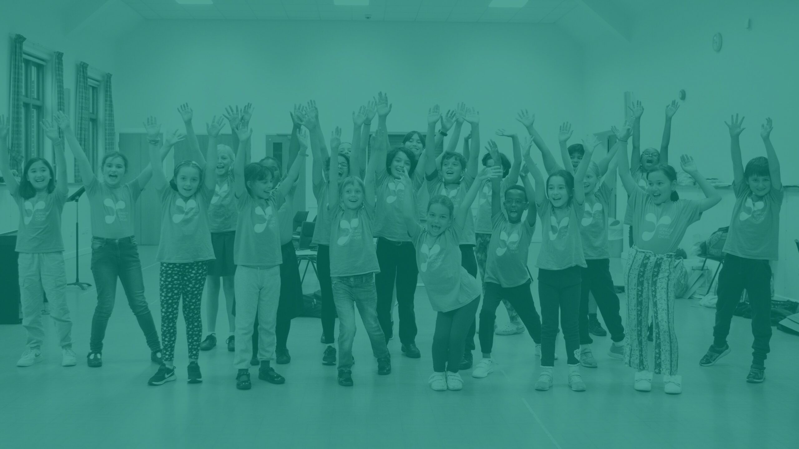 A teal-washed image of a group of 20 children wearing LYC T shirts, all with both hands up at the air and smiling at the camera. They are in a rehearsal room.